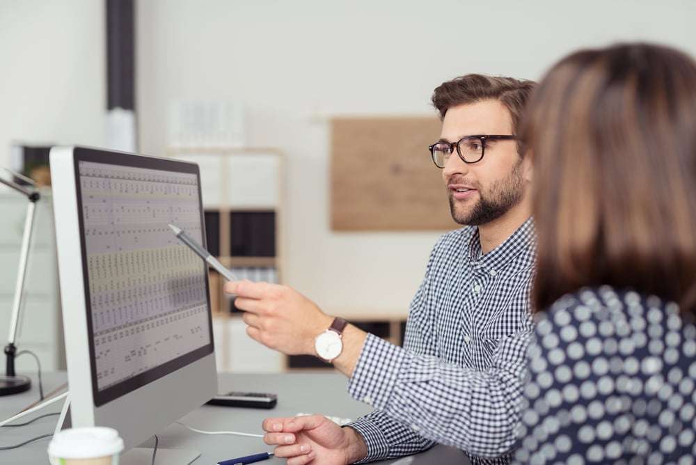 Proficient young male employee with eyeglasses and checkered shirt, explaining a business analysis displayed on the monitor of a desktop PC to his female colleague, in the interior of a modern office